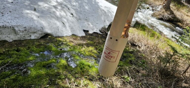 A copper water bottle sits on patchy snow along a forest trail with evergreen trees and a stream in the background