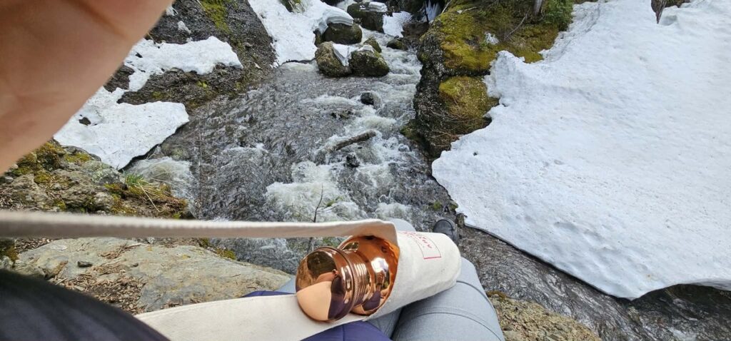 Copper Moscow mule mug held over a rocky snow-lined mountain stream during a hike