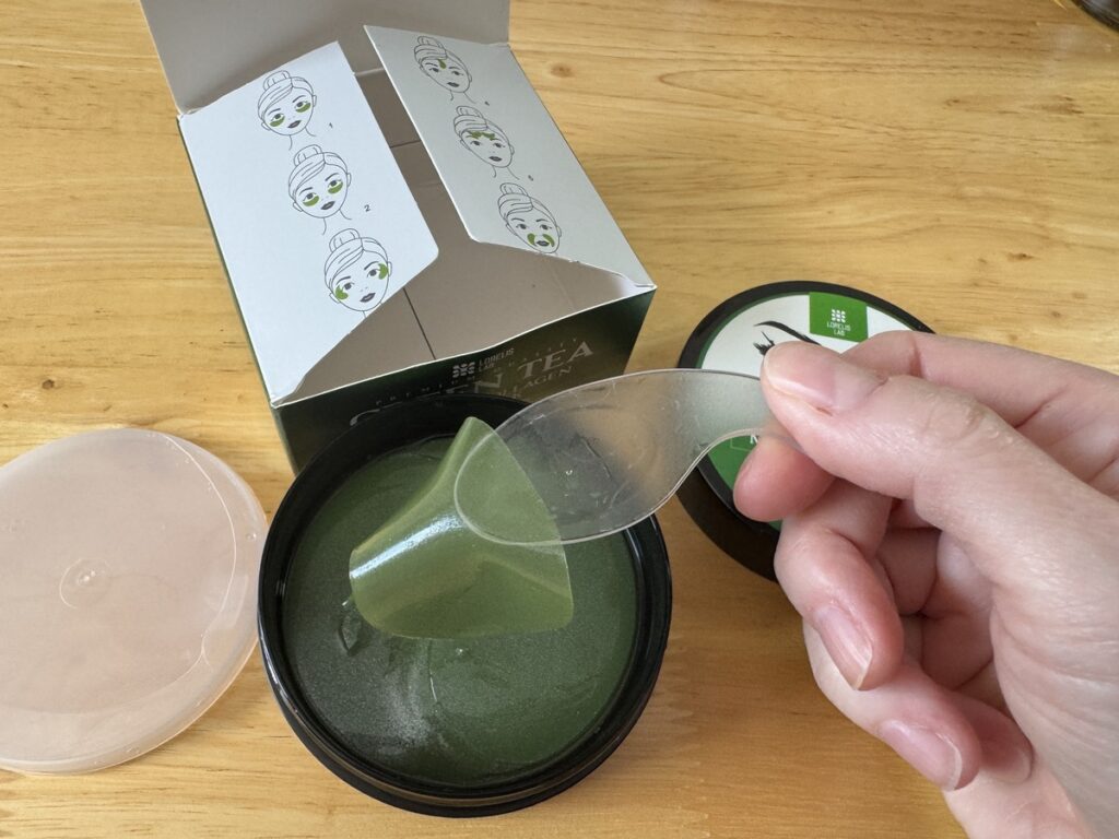 Hand holding a clear spatula scooping green tea hydrogel eye patches from an open jar beside the product box