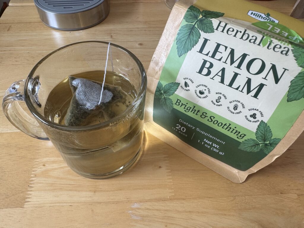 Lemon Balm Herbal Tea bag steeping in a clear glass mug next to the green Bright and Soothing pouch on a wood table