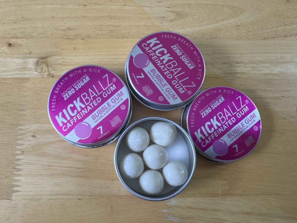 Three tins of KickBallz Caffeinated Bubble Gum on a wood surface, one open showing white gum balls inside