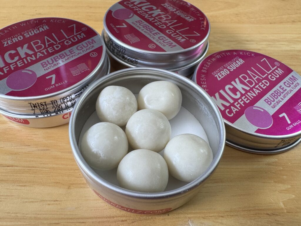 Three pink KickBallz Caffeinated Bubble Gum tins on a wood surface with one open tin showing white gum balls inside