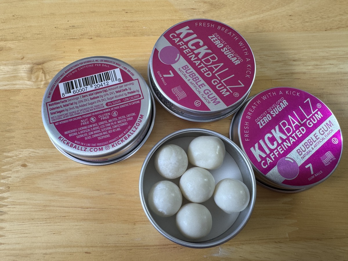 Three KickBallz Caffeinated Bubble Gum tins on a wood surface, one open showing white gum balls inside