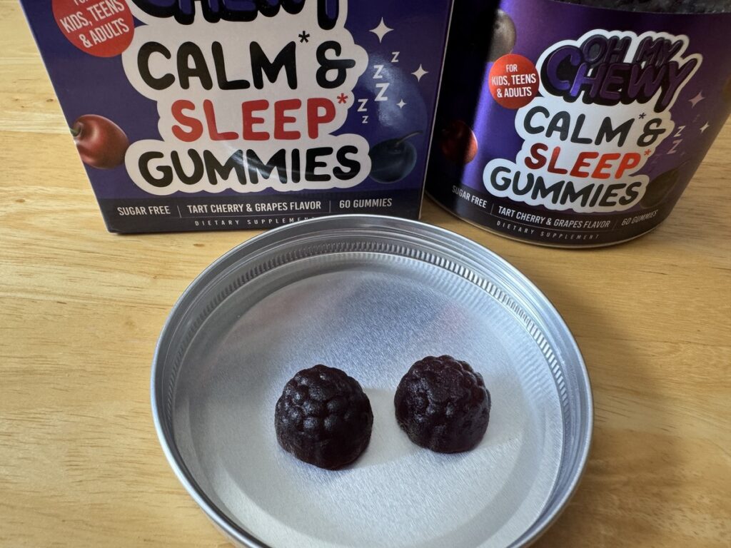 Open jar of Oh My Chewy dark berry-shaped gummies next to its purple branded box and metal lid on a wood surface