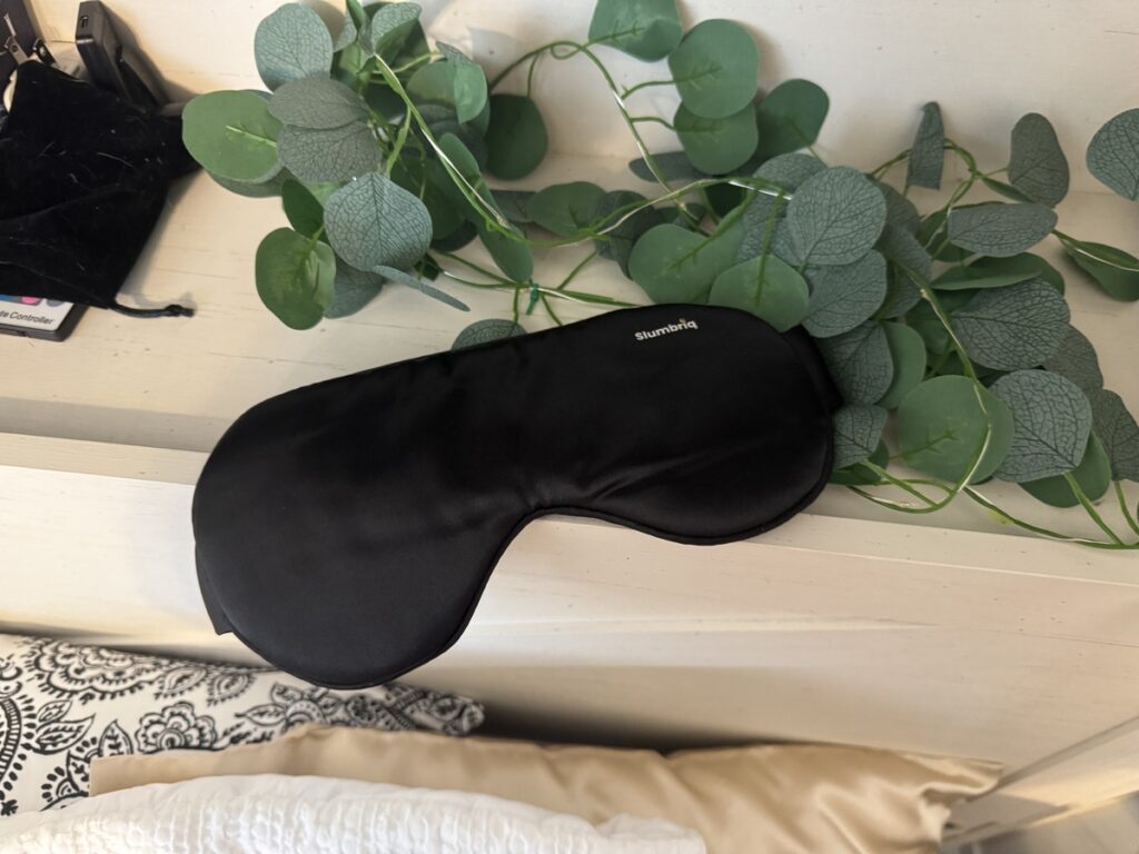Black Slumbriq sleep mask resting on a white wood surface with eucalyptus sprigs and a velvet pouch