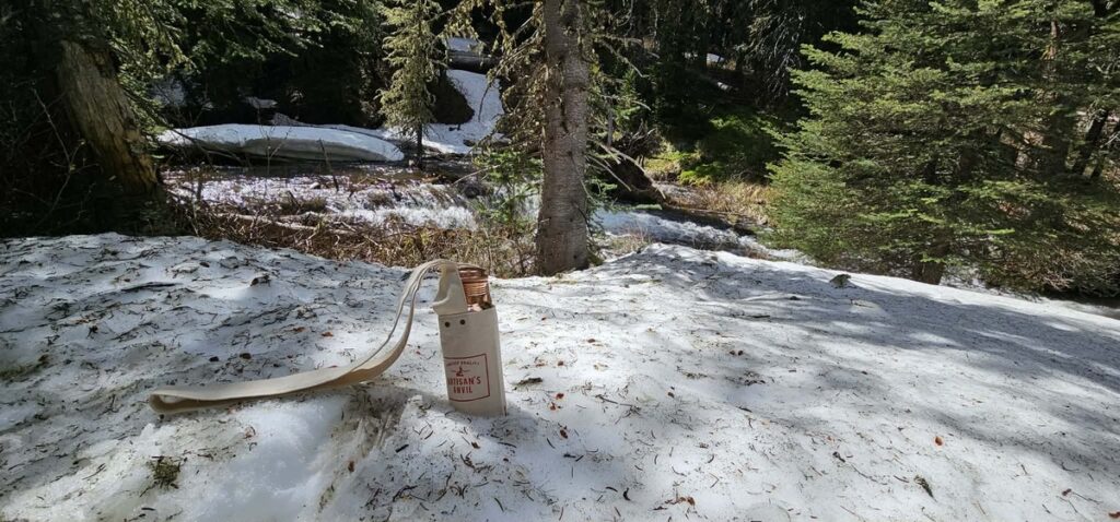 A water filter bottle stands upright on a patch of snow beside a small creek and evergreen trees in a forest