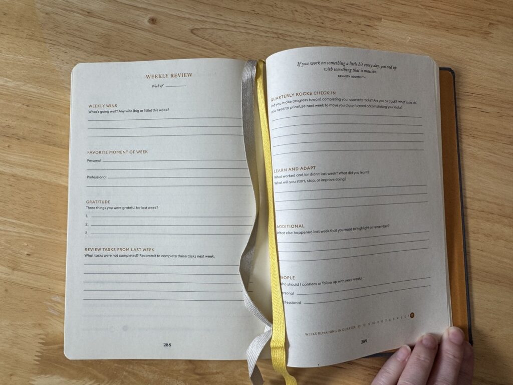 Open journal on a wood table showing a Weekly Review spread with gratitude and reflection prompts and a yellow ribbon