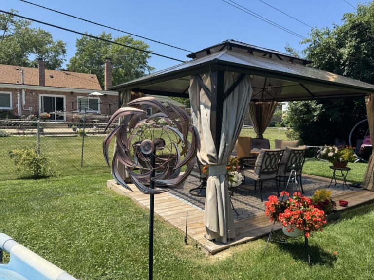 Backyard scene with a hardtop gazebo, patio furniture, red flowers, and a copper kinetic wind spinner sculpture on the lawn.