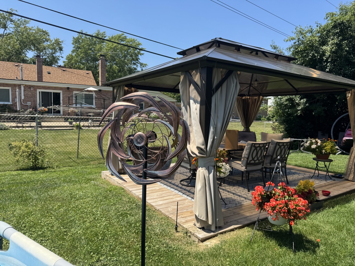 Backyard scene with a hardtop gazebo, patio furniture, red flowers, and a copper kinetic wind spinner sculpture on the lawn.