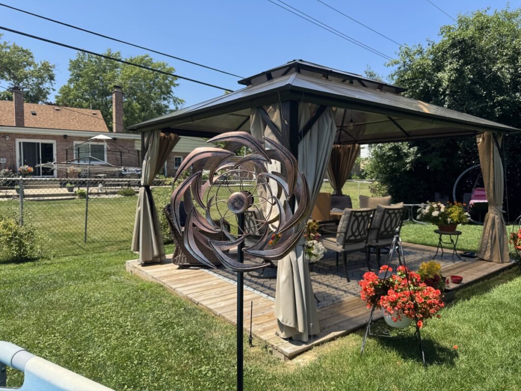 Backyard with brown hardtop gazebo, patio furniture, red geraniums, and a large copper kinetic wind spinner sculpture.