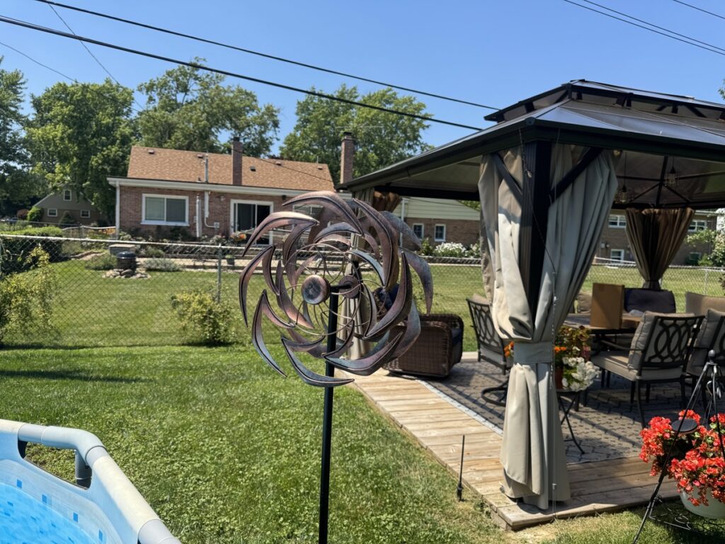 Backyard with copper kinetic wind spinner, curtained gazebo with patio furniture, pool edge, and brick house in background.