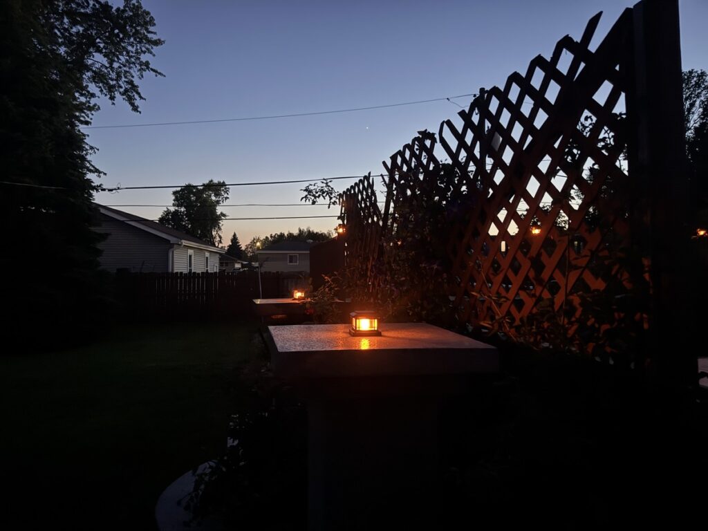 Glowing flame-effect lantern on a stone pillar in a backyard at dusk with a wooden lattice trellis behind it.