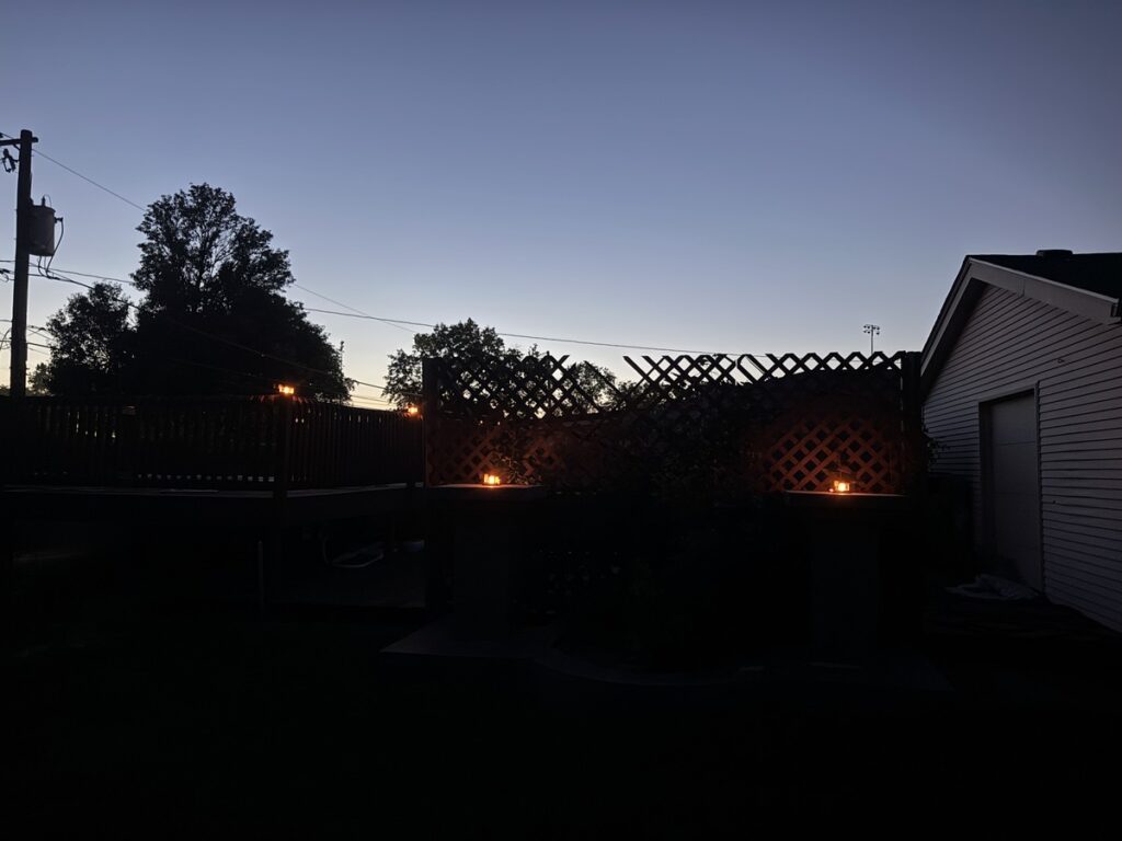 Backyard at dusk with wooden lattice fence, glowing lanterns, silhouetted trees, and a house under a twilight sky.