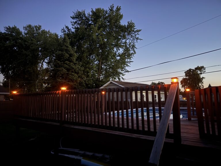 Backyard wooden fence around an in-ground pool at dusk with warm string lights glowing and a deep blue sky.