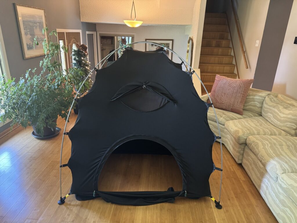Black dome pop-up privacy tent with a peekaboo window on hardwood floor in a living room beside a striped sofa.