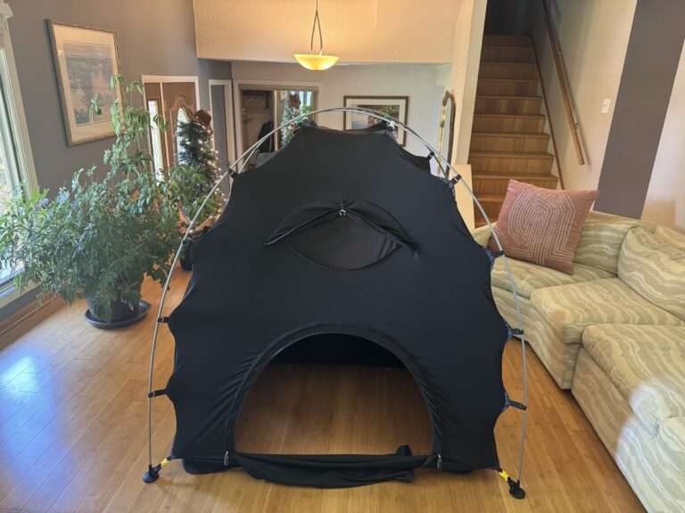 Black dome-shaped blackout canopy bed tent set up on a hardwood floor in a living room with a striped green couch.