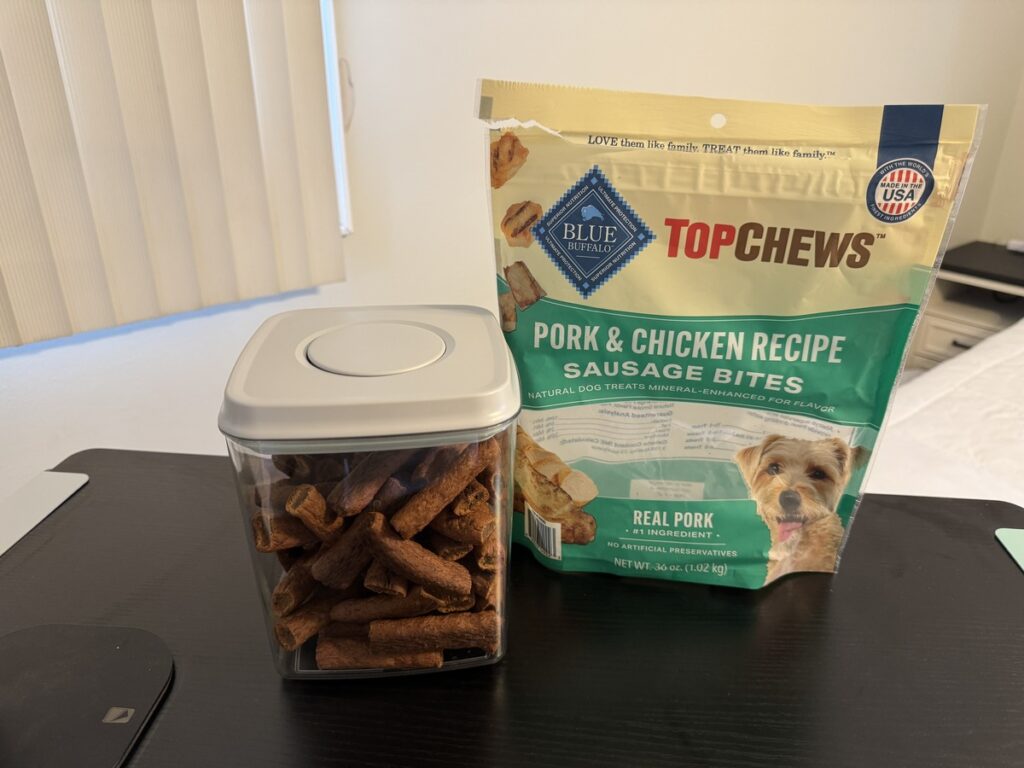 Blue Buffalo Top Chews Pork and Chicken Recipe Sausage Bites bag beside a clear storage container filled with the treats.