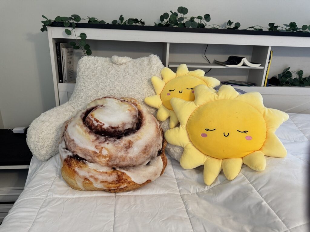 A giant cinnamon roll plush pillow and two smiling yellow sun plush pillows on a white bed with a sheepskin cushion.