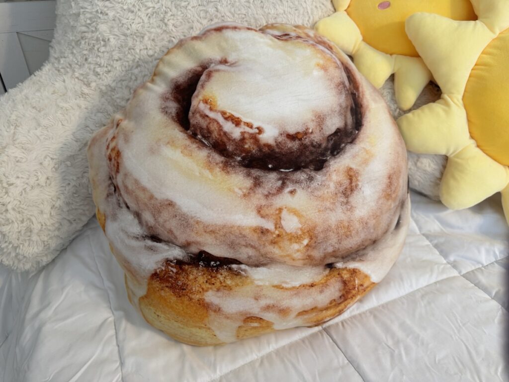 Large cinnamon roll shaped plush pillow with icing print on a white bed next to a yellow sun plush.