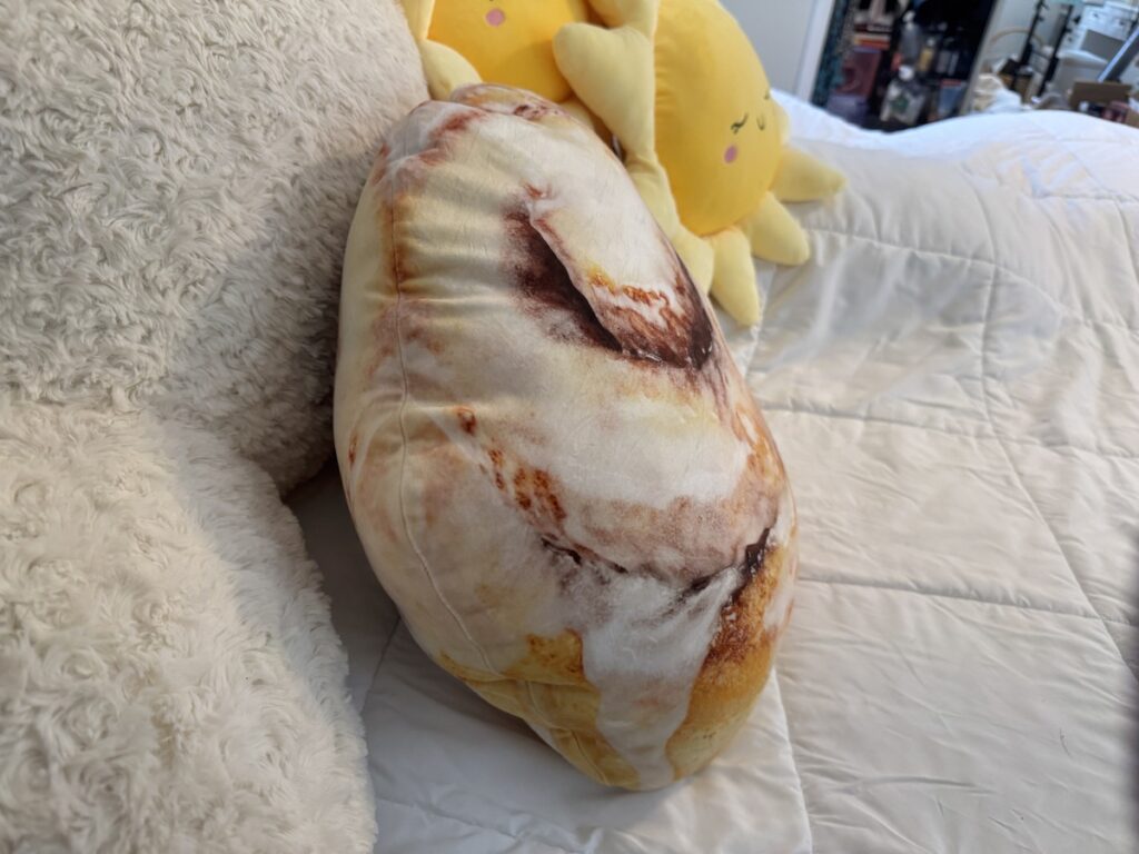 Large cinnamon roll shaped plush pillow resting on a white bed next to a smiling yellow sun plush.