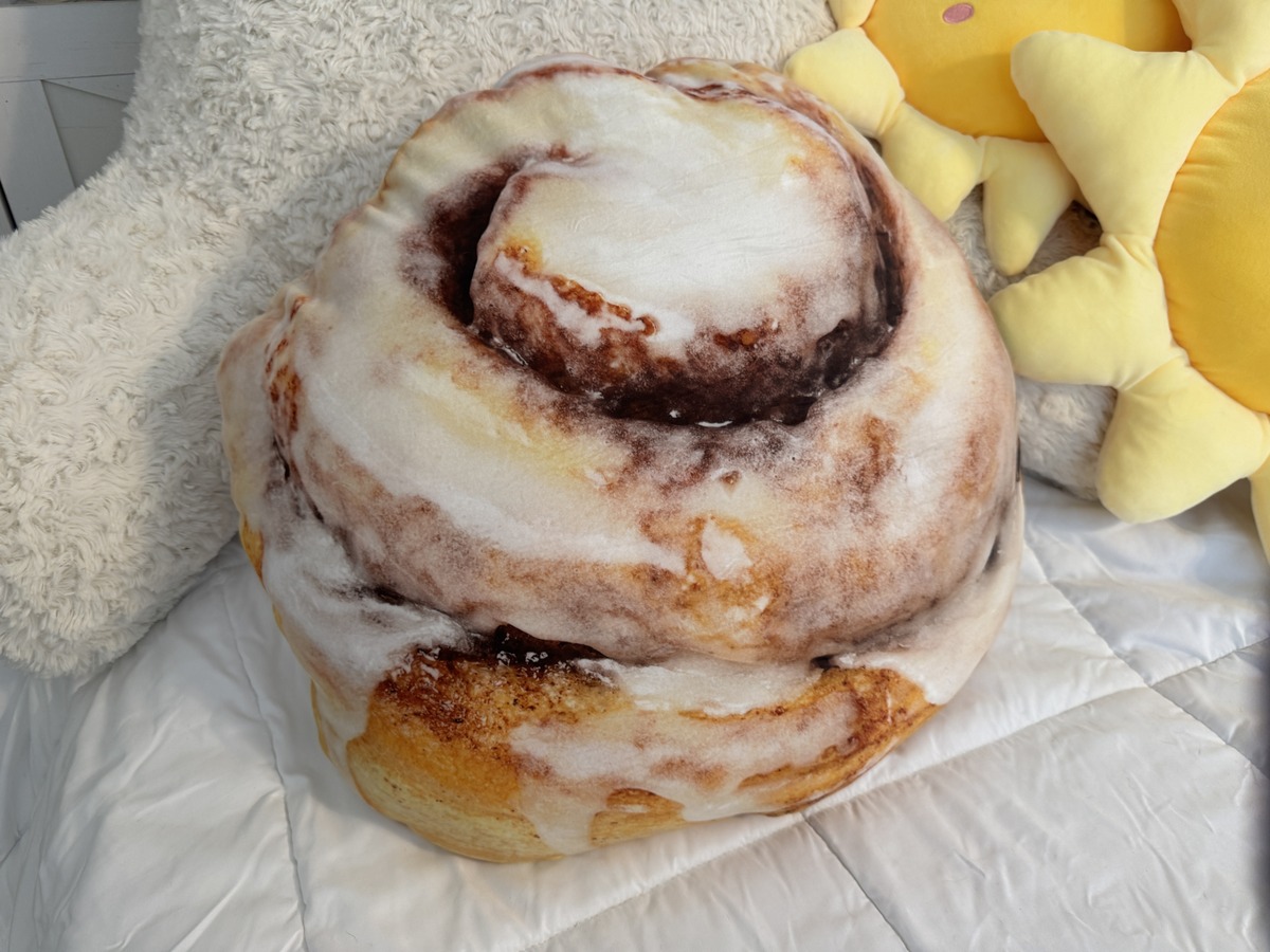 Large cinnamon roll shaped plush pillow with icing print on a white bed next to a yellow sun plush.