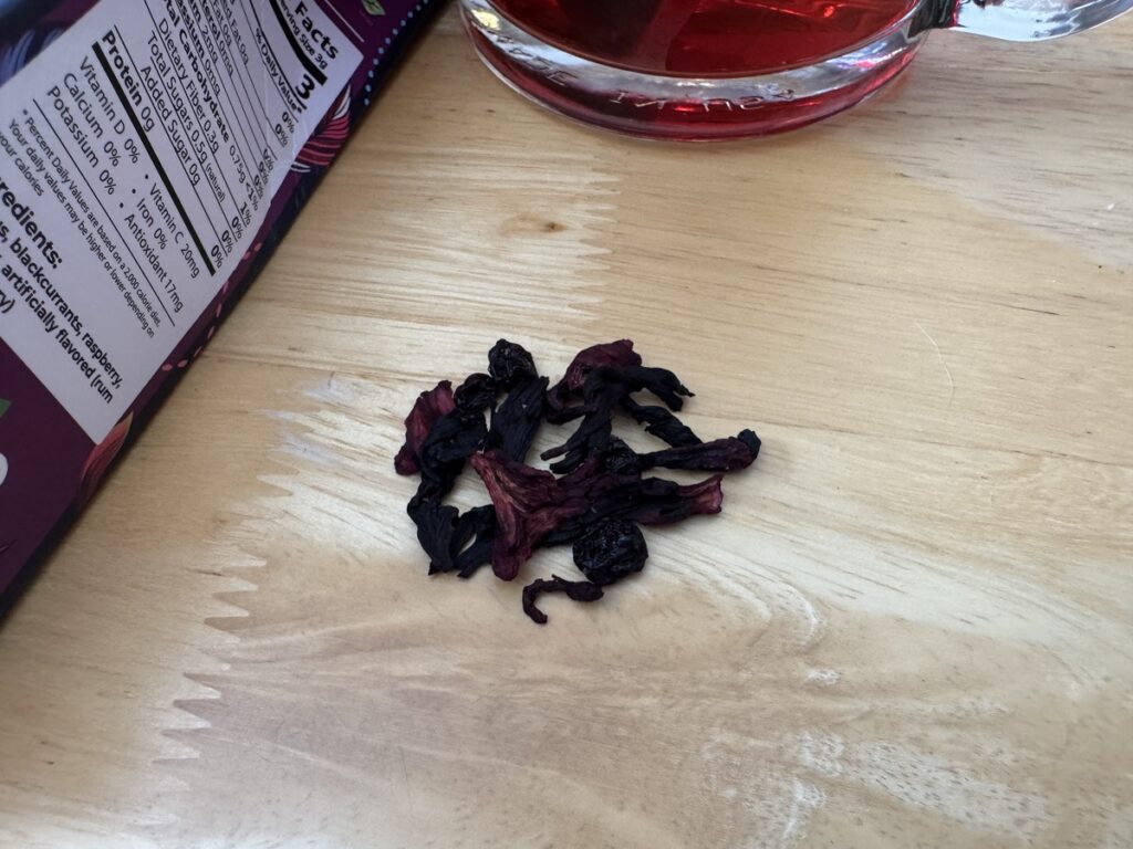 Pile of dried hibiscus petals on a light wood surface next to a glass of red hibiscus tea and a bag of dried flowers.