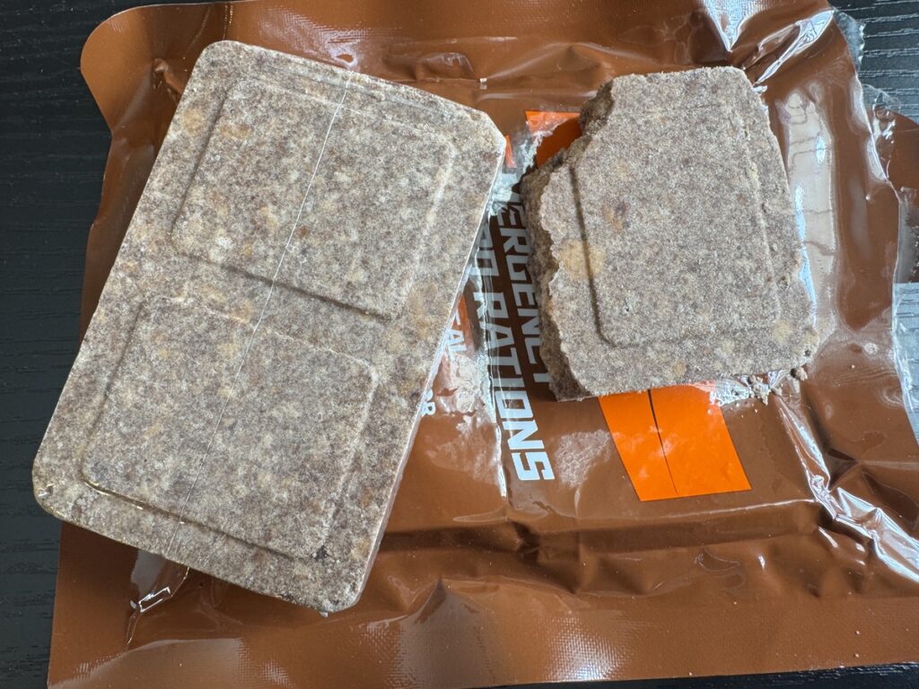 Two compressed brown ration bars resting on the torn orange Emergency Rations foil pouch they came from.