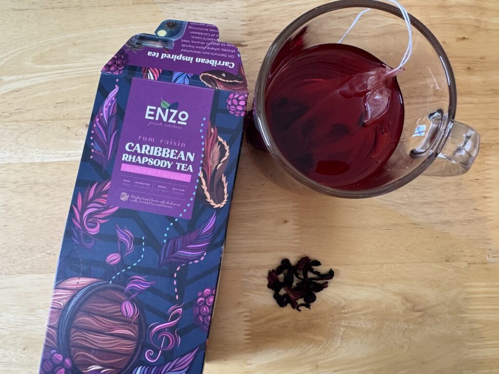 ENZO Caribbean Rhapsody rum raisin tea box beside a glass mug of brewed deep red hibiscus tea and loose petals on wood.