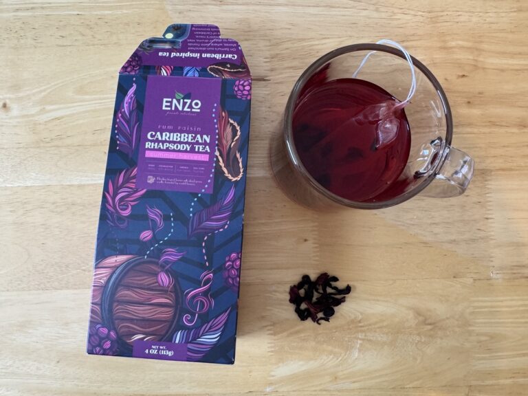 Enzo Caribbean Rhapsody rum raisin tea carton beside a glass mug of brewed deep red hibiscus tea with loose leaves.