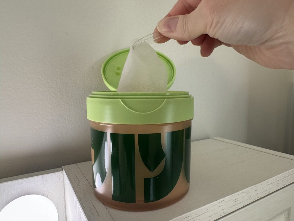 Hand pulling a white wet wipe from a green and gold canister with a bamboo pattern on a white dresser.