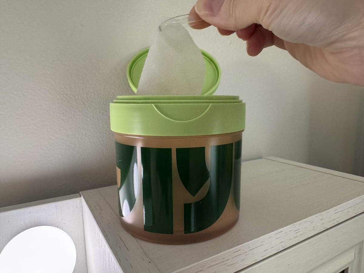 Hand pulling a wipe from a green and bronze bamboo-print canister sitting on a white dresser.