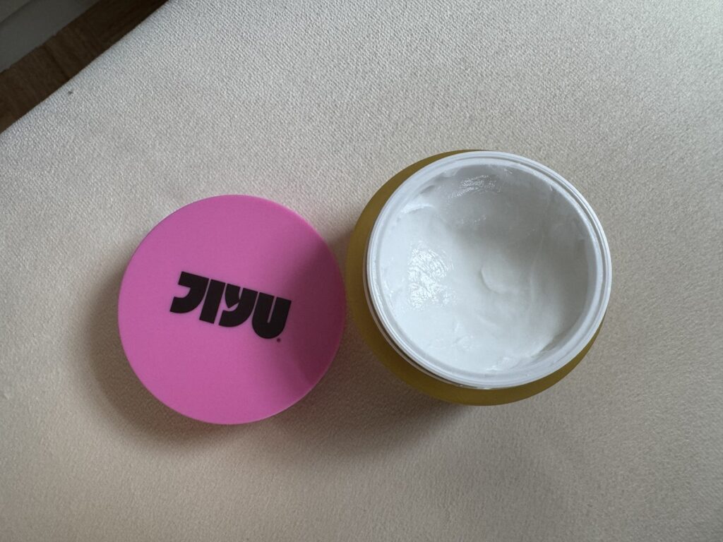 Open jar of Jiyu body butter with pink lid off to the side showing white cream inside a mustard yellow container.