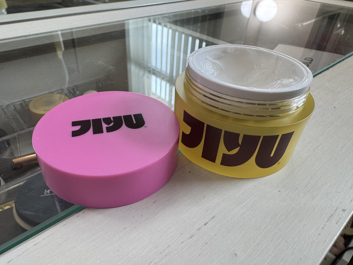 Two Jiyu hair pomade jars on a glass shelf, a closed pink jar and an open yellow jar showing white cream inside.