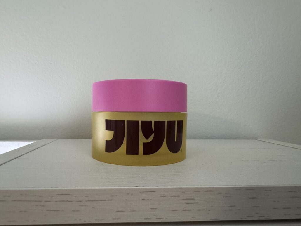 Small round Jiyu jar with pink lid and gold body featuring stylized brown lettering, sitting on a white shelf.