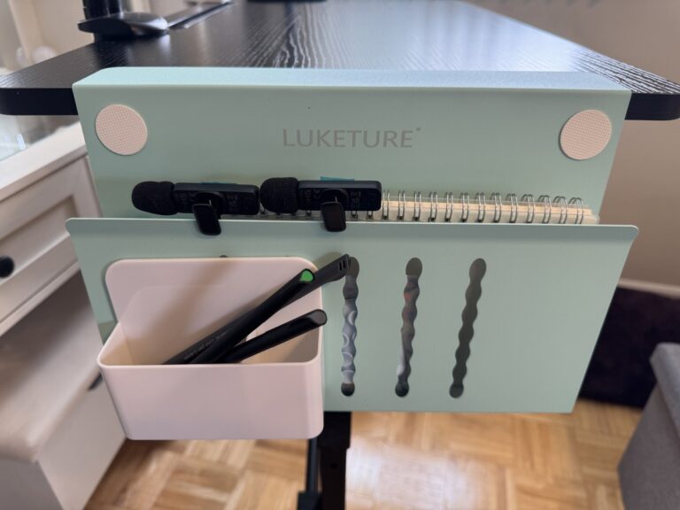 Mint green LUKETURE under-desk clip-on organizer holding a spiral notebook, binder clips, and pens on a black desk.