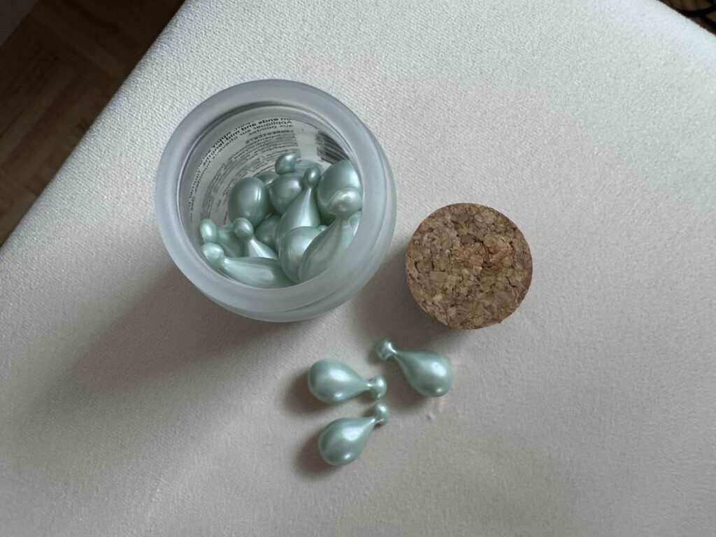 Frosted glass jar with cork lid holding mint green Remilia Cosmocap hair serum capsules on cream fabric.