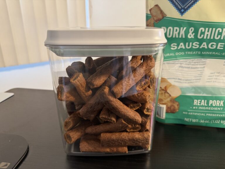 Clear storage container filled with Pork and Chicken Sausage dog treats next to the green 36 oz bag.