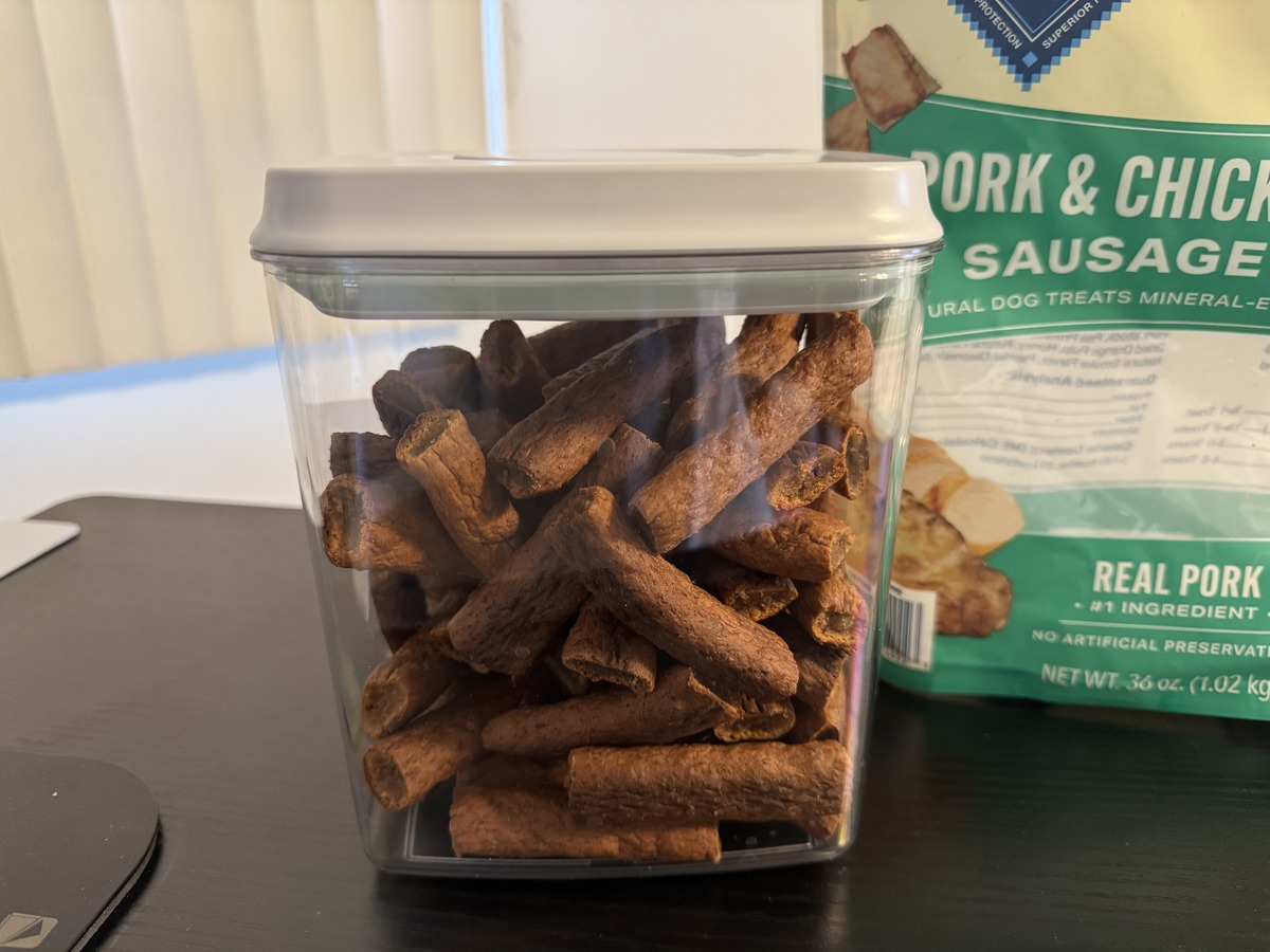 Clear storage container filled with Pork and Chicken Sausage dog treats next to the green 36 oz bag.
