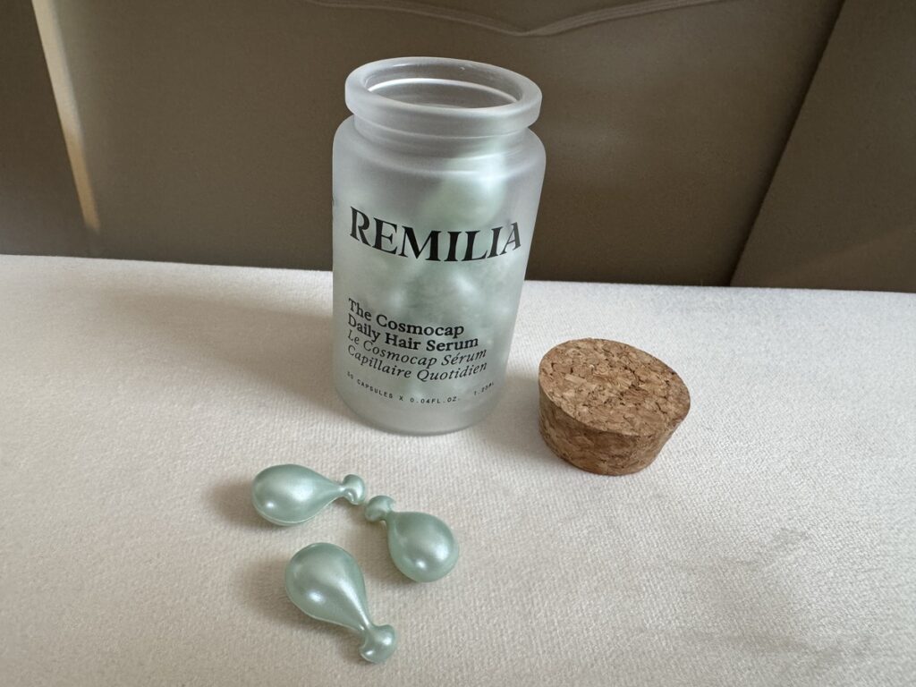 Frosted glass Remilia The Cosmocap Daily Hair Serum bottle with cork stopper and three pearlescent green serum capsules.