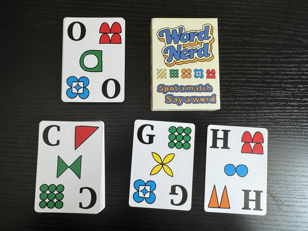 Word Nerd card game box with four letter cards showing O, C, G, and H spread on a dark wood table.