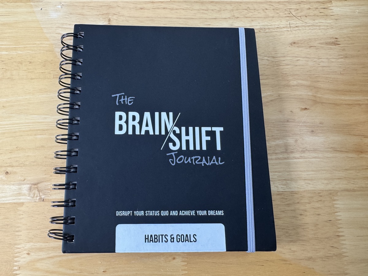 The Brain Shift Journal Habits and Goals edition, a black spiral-bound notebook resting on a wood table.