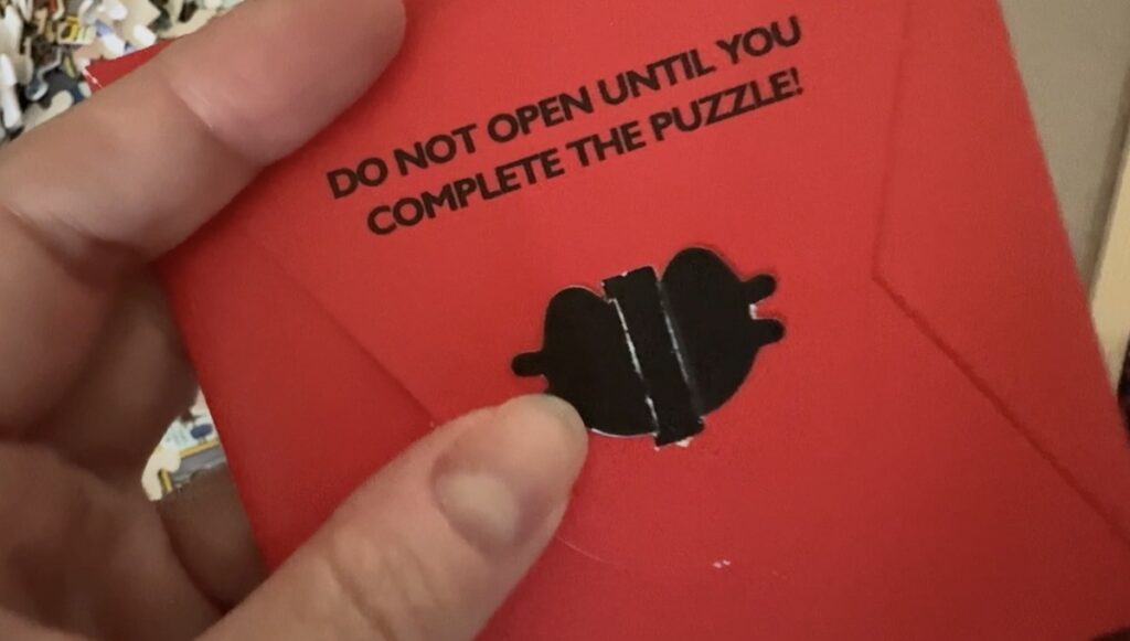 Hand holding a red puzzle envelope printed with Do Not Open Until You Complete The Puzzle and a black seal.