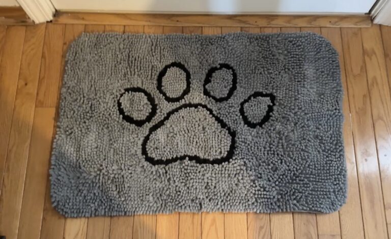Gray chenille shag doormat with a black paw print design laid on a hardwood floor in a doorway.