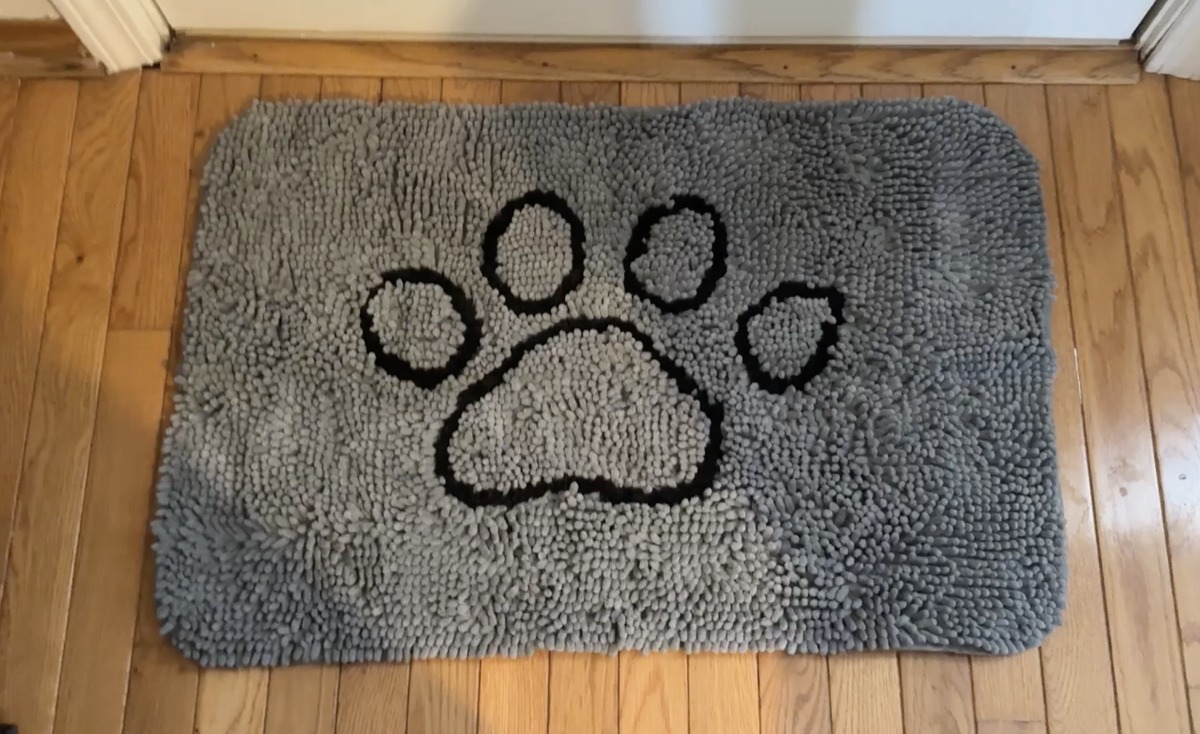 Gray chenille shag doormat with a black paw print design laid on a hardwood floor in a doorway.