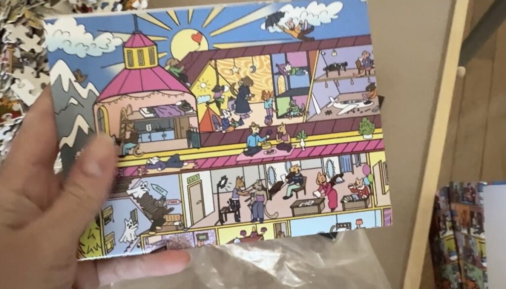 Hand holds a colorful illustrated hidden-picture puzzle showing a cutaway building with cartoon characters in various rooms.