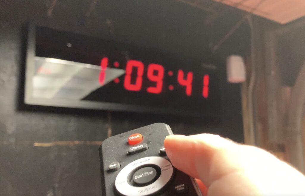 Hand holding a small black remote with a Clock button in front of a wall-mounted digital clock reading 1:09:41 in red.