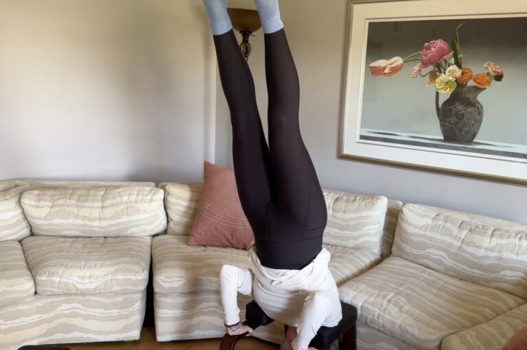 Person doing a headstand on a wooden stool in a living room with a beige striped sectional and framed floral still-life.