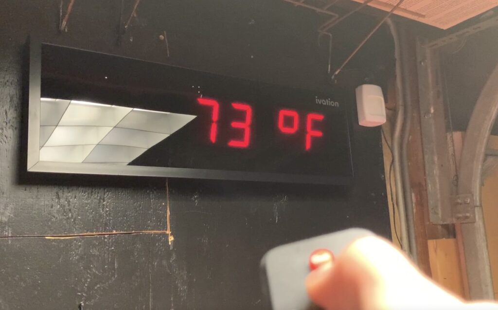 Ivation wall-mounted LED thermometer showing 73 degrees Fahrenheit in red digits, with a hand holding a remote below.