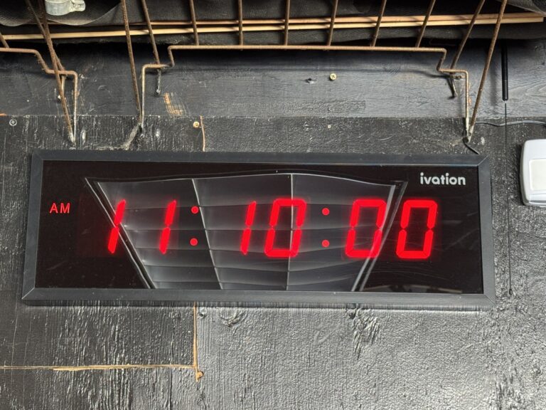 Ivation digital LED wall clock showing 11:10:00 AM in red numerals, mounted on a black wire shelving unit.