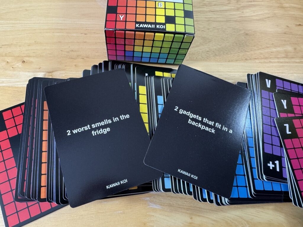 Spit Out First card game spread with the Kawaii Koi box, black prompt cards and colorful grid-letter cards.